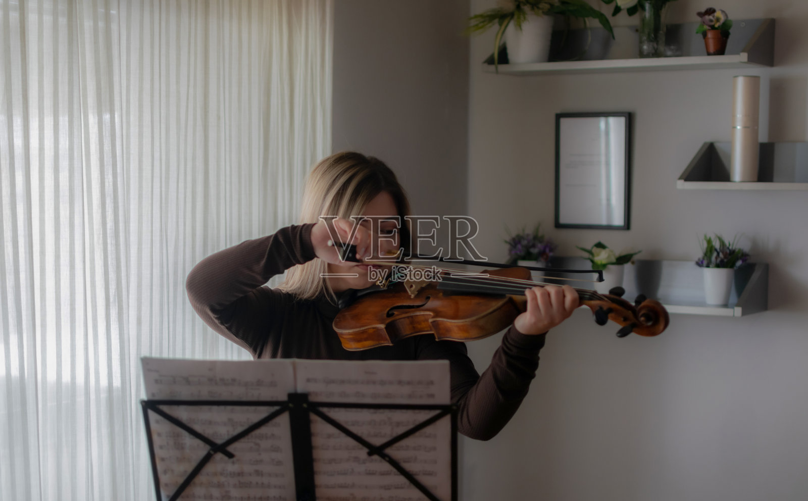 美丽和年轻的女孩享受在她的家里演奏她的中提琴，练习和享受她的自由时间，在阳光明媚的日子里在窗户附近演奏。照片摄影图片