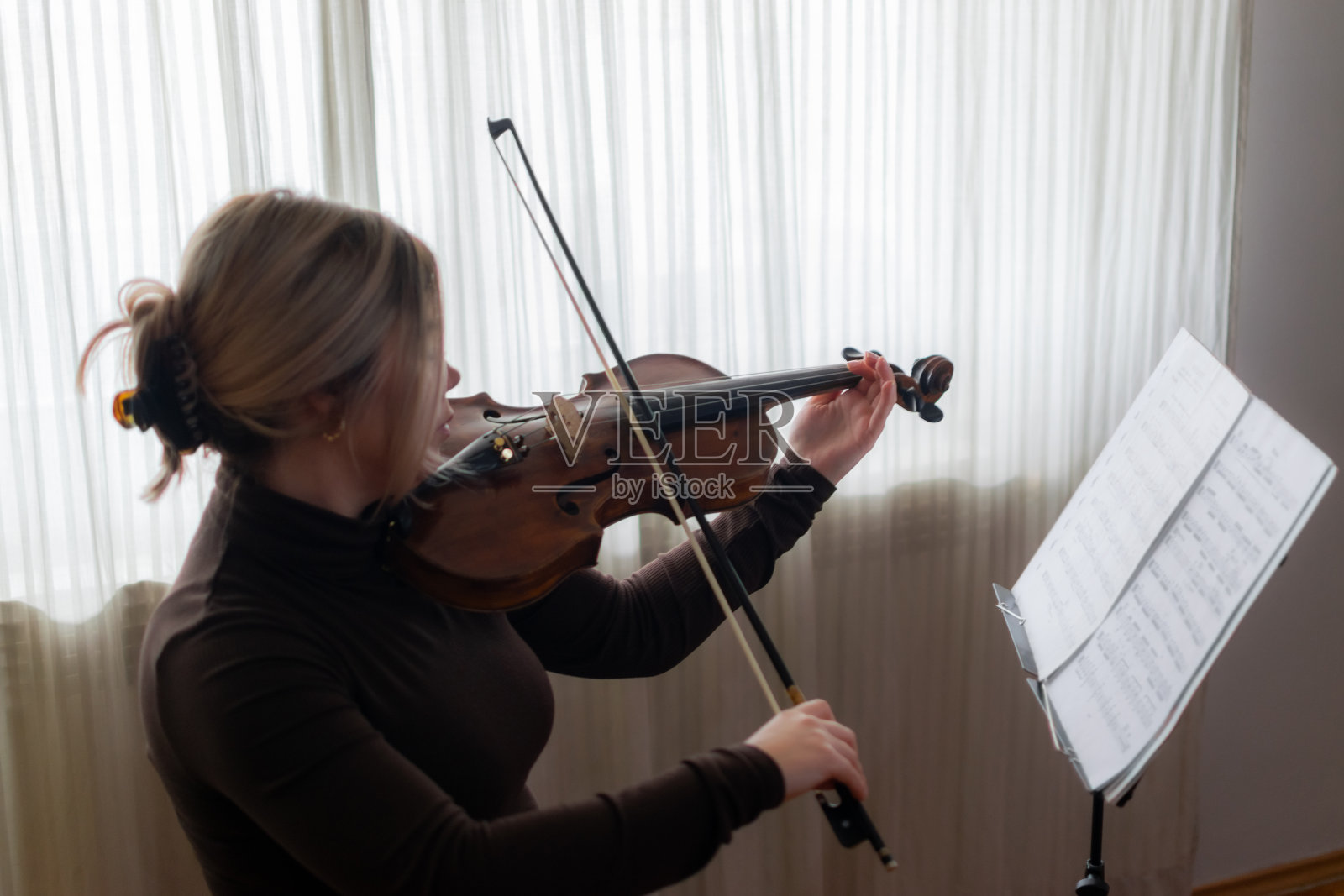 美丽和年轻的女孩享受在她的家里演奏她的中提琴，练习和享受她的自由时间，在阳光明媚的日子里在窗户附近演奏。照片摄影图片