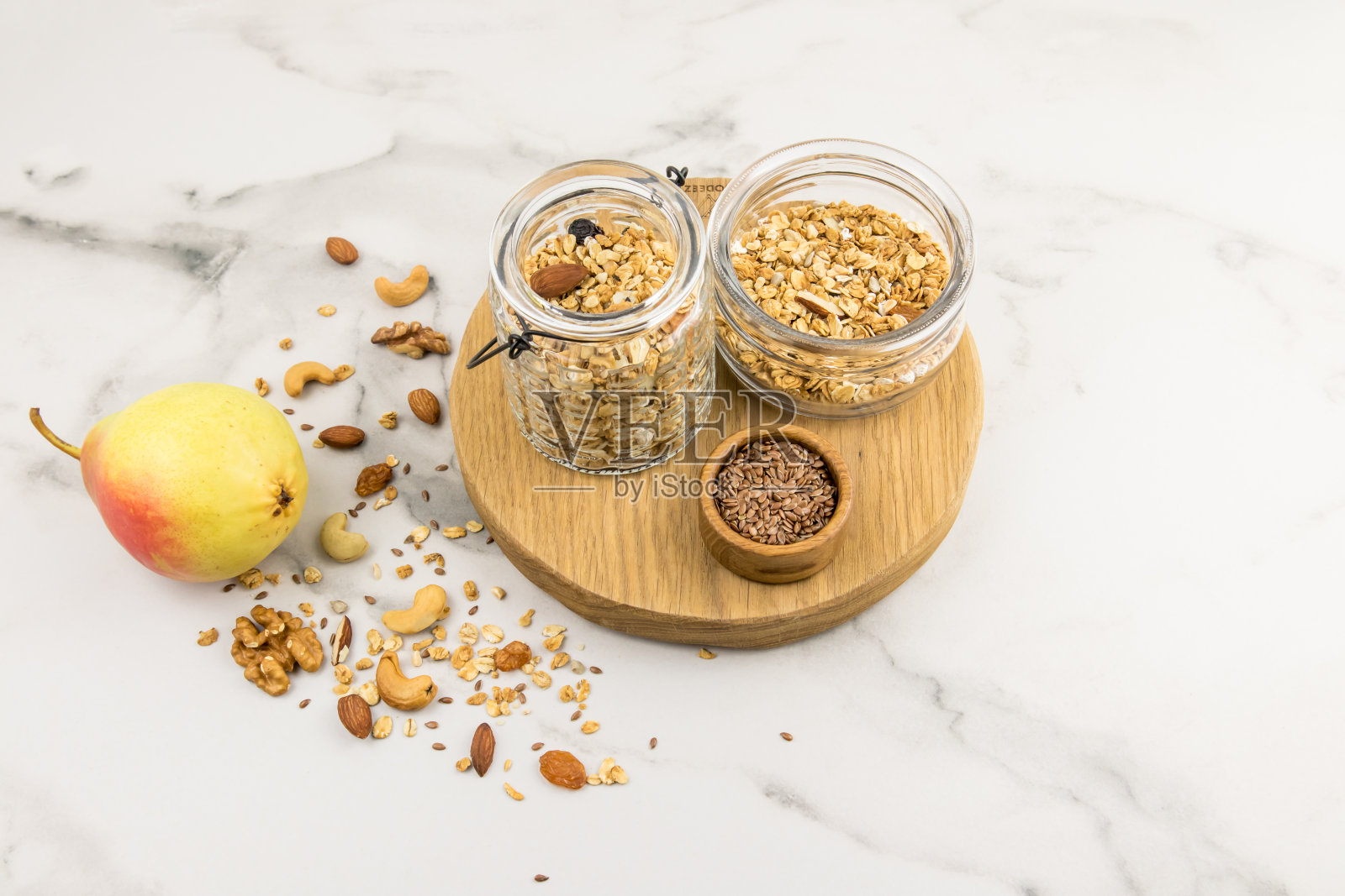 两个不同的玻璃罐与核桃格兰诺拉站在切割圆板上。大理石背景，上面散落着坚果和熟梨。照片摄影图片