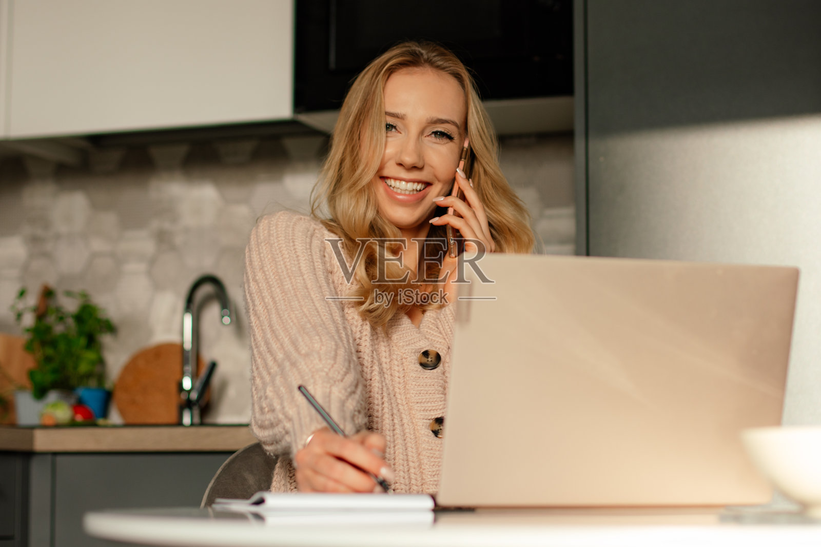 金发微笑的年轻女子工作在笔记本电脑和电话。女性在大流行中远程工作。照片摄影图片