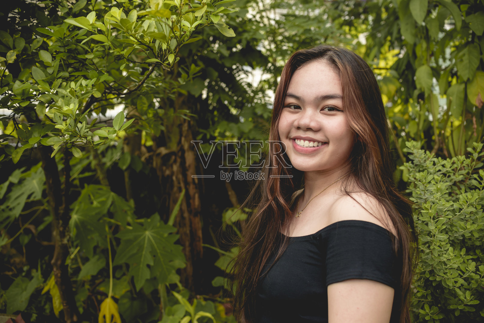 一个年轻、漂亮的十几岁亚洲女孩对着镜头微笑。郁郁葱葱的夏日绿叶背景。照片摄影图片