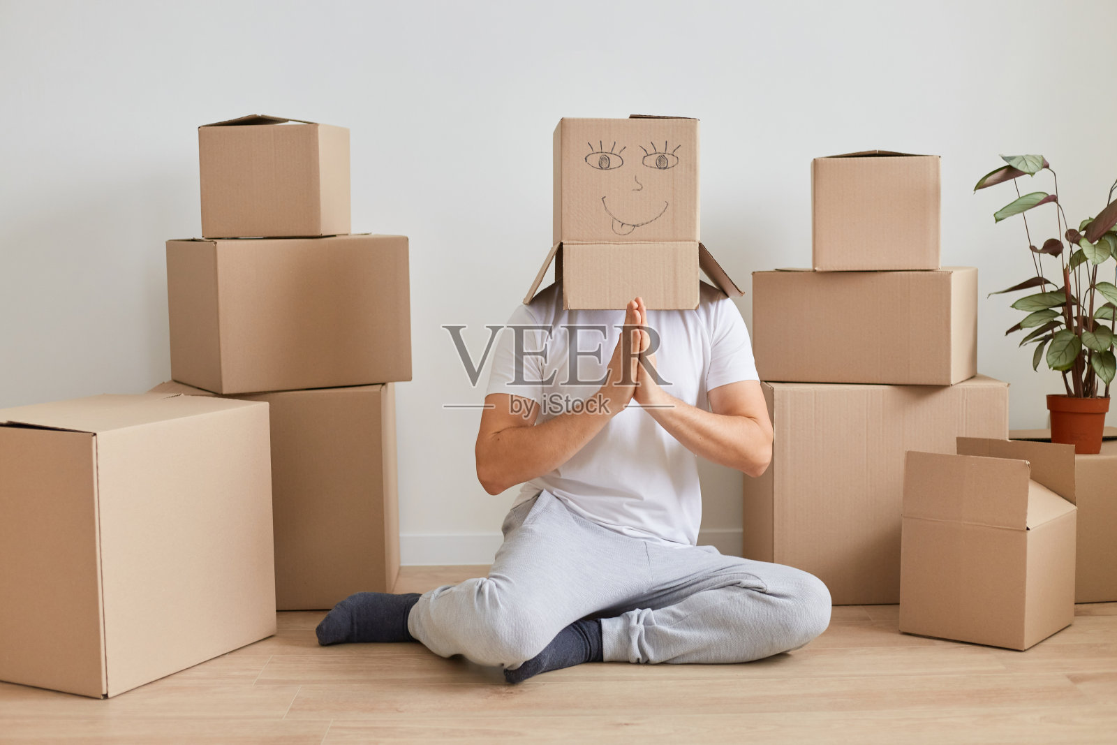 在室内拍摄的这张照片中，一名不知名的男子穿着白色t恤，坐在纸板箱旁边的地板上，把纸板箱顶在头上摆姿势，在搬新家期间，他试图通过瑜伽练习放松自己。照片摄影图片