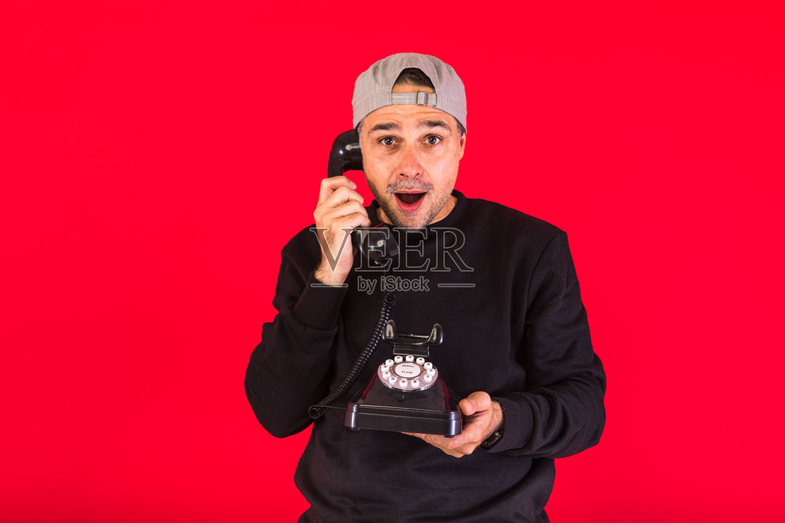 一名男子戴着一顶黑色的帽子，用一部60年代的复古电话接了一个电话，他惊讶地发现，电话背景是红色的，上面有复印空间。接触、社会关系和电话的概念照片摄影图片