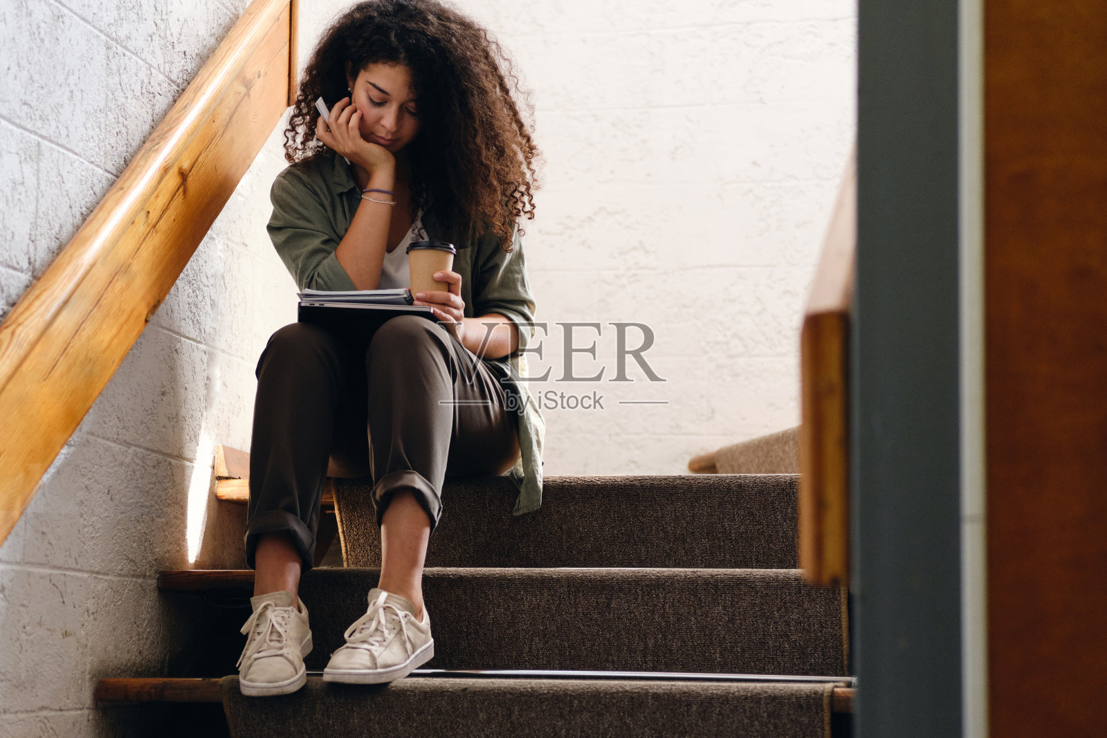 一头黑色卷发的年轻迷人的女学生坐在大学的楼梯上，膝盖上放着课本，手里拿着一杯咖啡，沉思地独自学习照片摄影图片