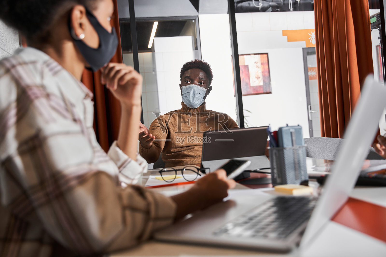 年轻的员工戴着防护口罩坐在办公室里，手里拿着电子产品，一边讨论照片摄影图片