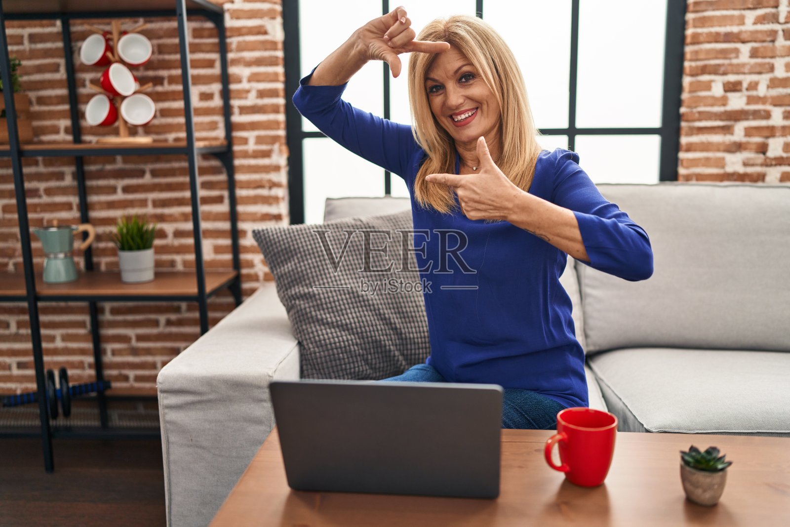 中年金发女人用电脑笔记本电脑在家里微笑着用手和手指做框架，快乐的脸。创意和摄影理念。照片摄影图片