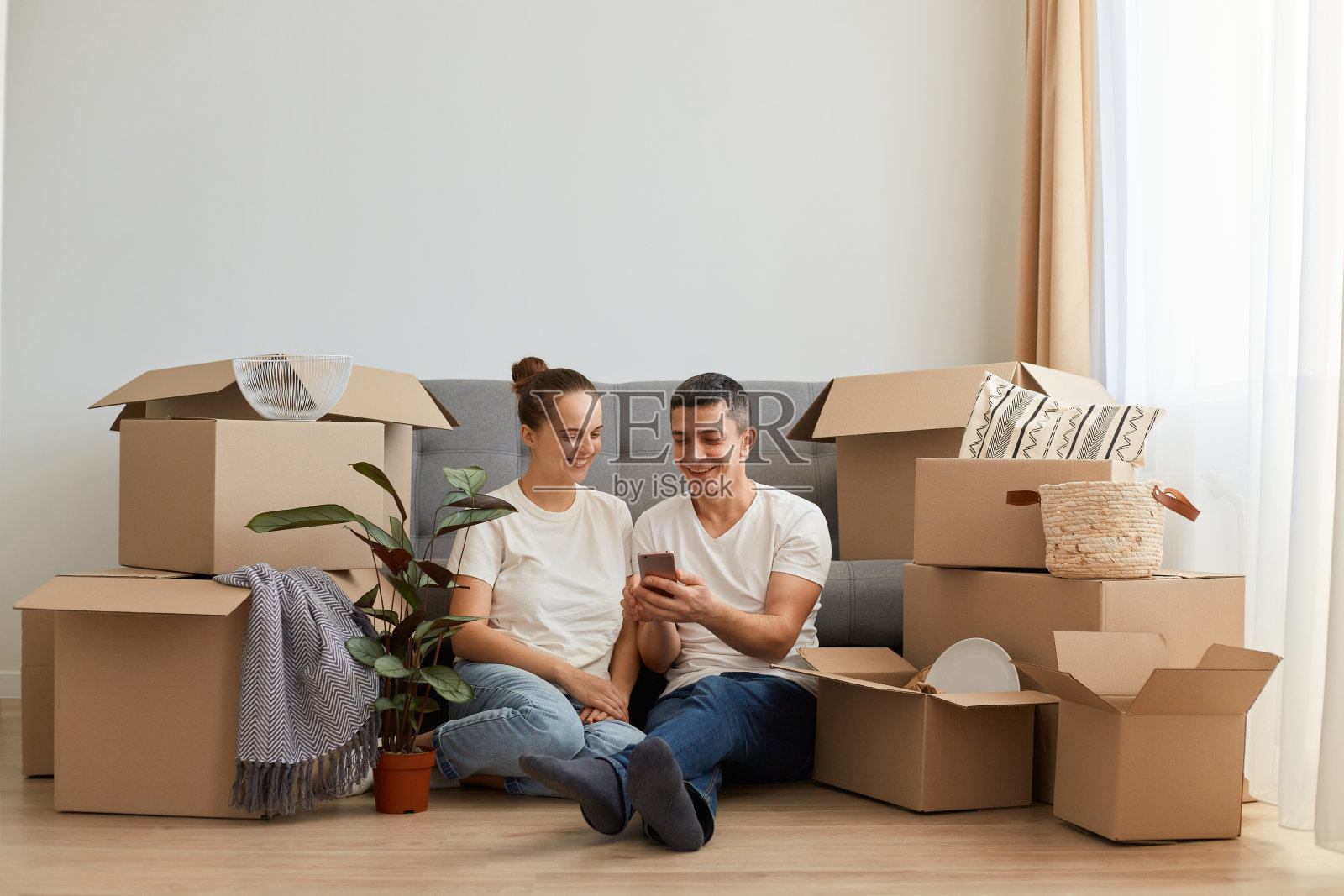 幸福的夫妻搬进新买的房子，在网上买家具，使用现代科技，男人通过笔记本电脑上网和打电话，坐在沙发旁边的地板上。照片摄影图片