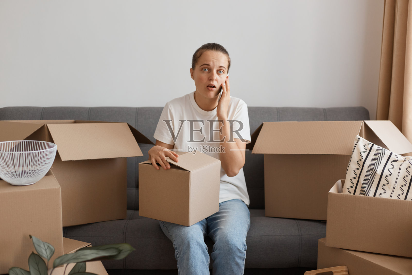 一名身穿白色t恤和牛仔裤的女子，在搬迁后的新公寓里，拿着包裹，坐在沙发上拍照，脸上露出惊讶的表情。照片摄影图片