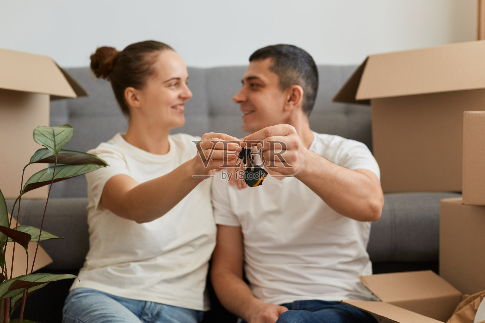 幸福积极的夫妇在新家，坐在沙发旁边的地板上，拿着他们新公寓的钥匙，用爱和温柔的目光看着对方，搬进新家。照片摄影图片