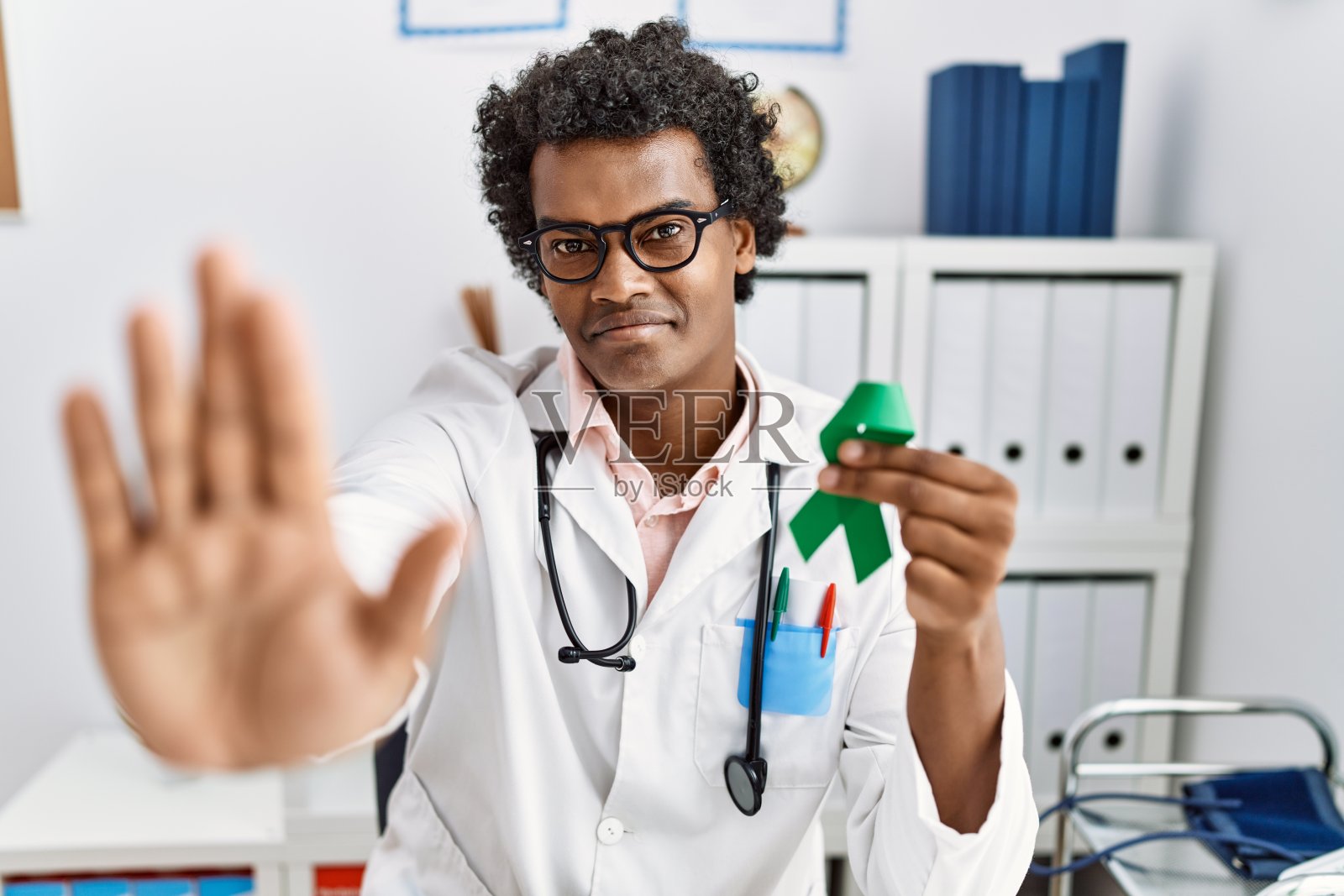 非洲医生手持支持绿丝带，张开手做停止标志，表情严肃自信，做防御手势照片摄影图片