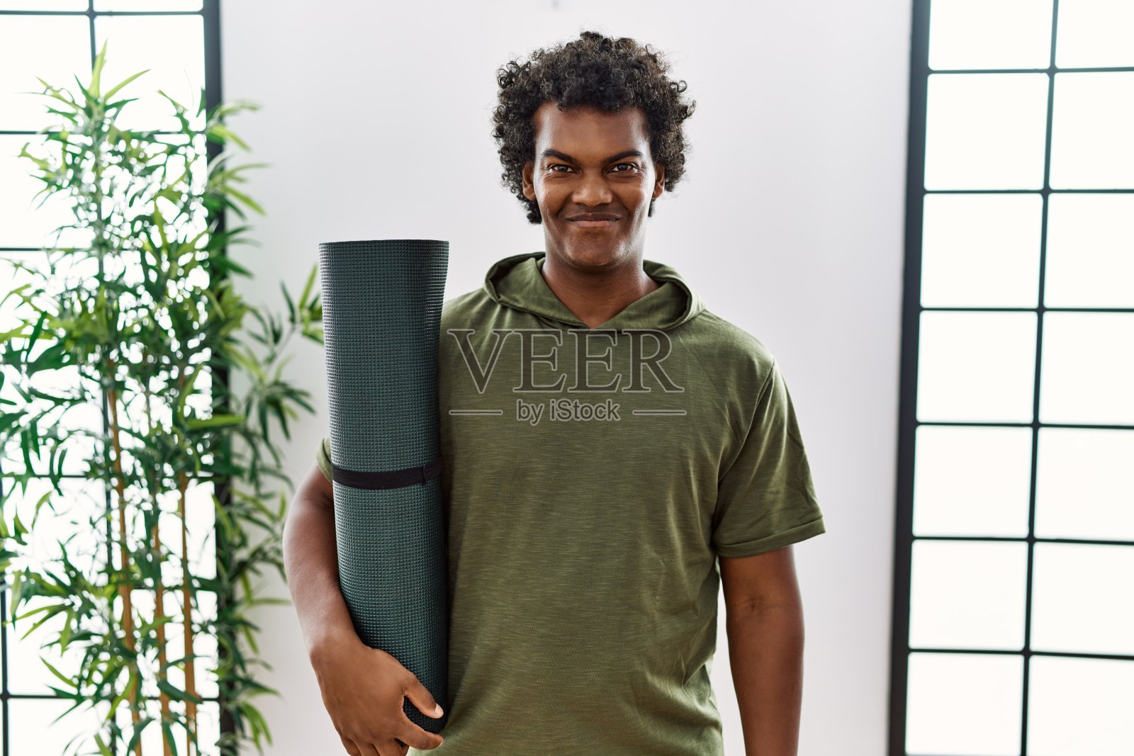 一头卷发的非洲男子拿着瑜伽垫在工作室里，脸上带着幸福和凉爽的微笑。幸运的人。照片摄影图片