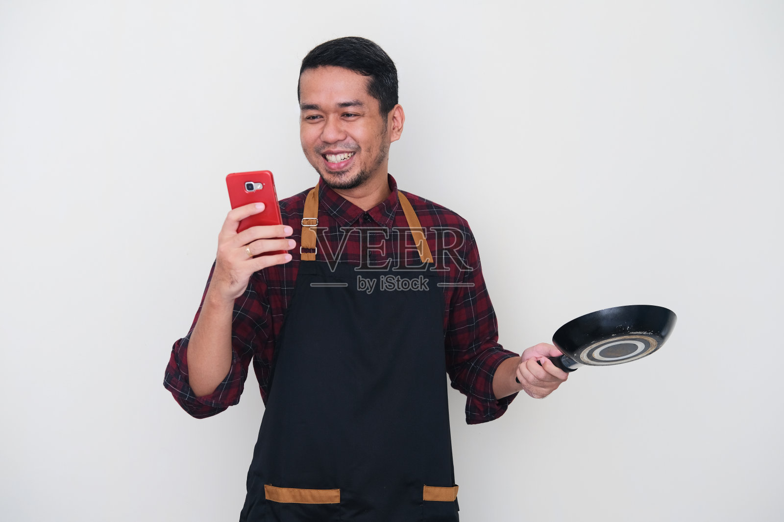 成年亚洲人穿着围裙微笑快乐时看他的手机和拿着小煎锅照片摄影图片