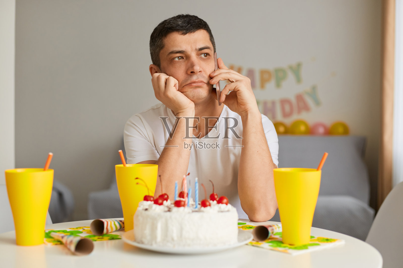 悲伤沉思的年轻人的肖像，穿着白色休闲风格的t恤，坐在桌子旁的蛋糕，与她的朋友谈论智能手机，独自庆祝生日，感到悲伤。照片摄影图片