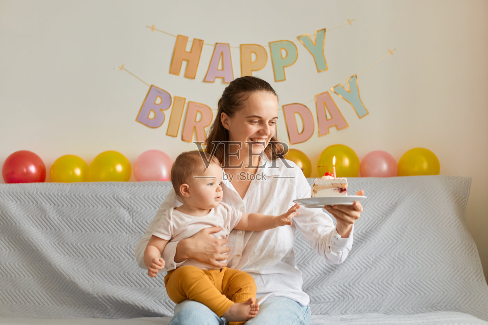 水平拍摄的快乐微笑的妇女坐在沙发上，她的婴儿女儿和拿着蜡烛的蛋糕，快乐地微笑，表达积极的情绪，庆祝第一个婴儿的生日。照片摄影图片