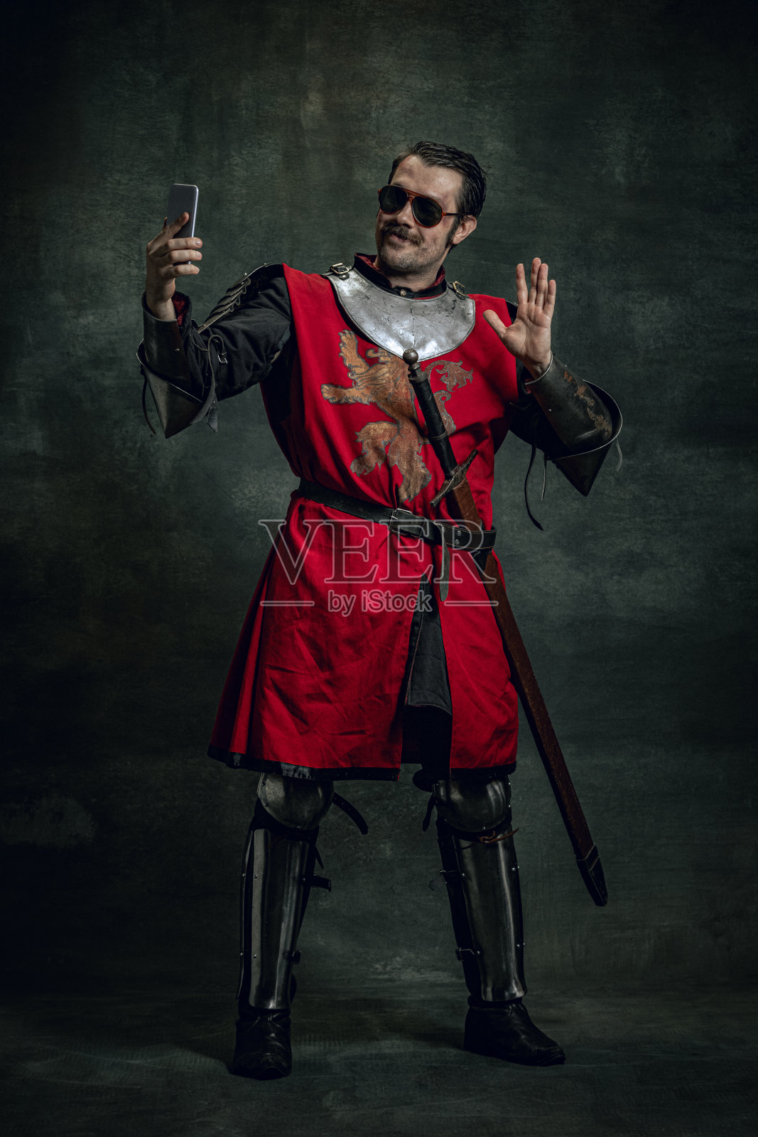 一个面带微笑的年轻男子，像中世纪的武士或骑士，脸上又脏又伤，在黑暗的背景下使用手机。时代的比较照片摄影图片