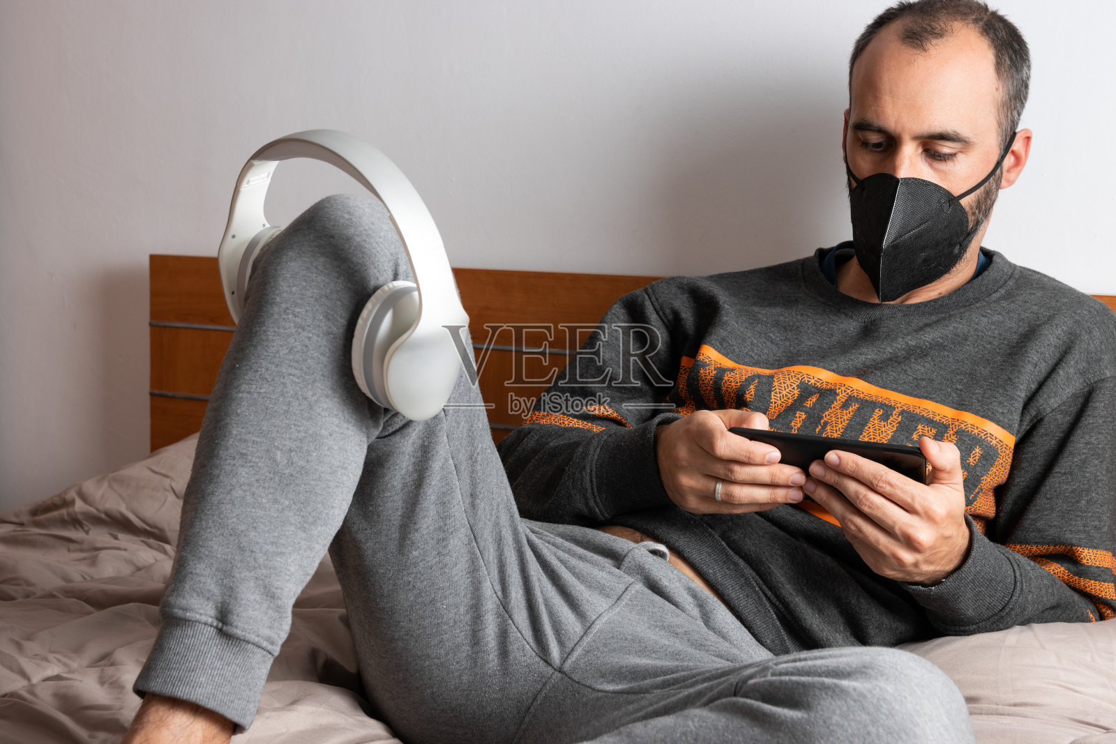 这名男子戴着耳机、手机和口罩，坐在床上，被限制在房间里照片摄影图片