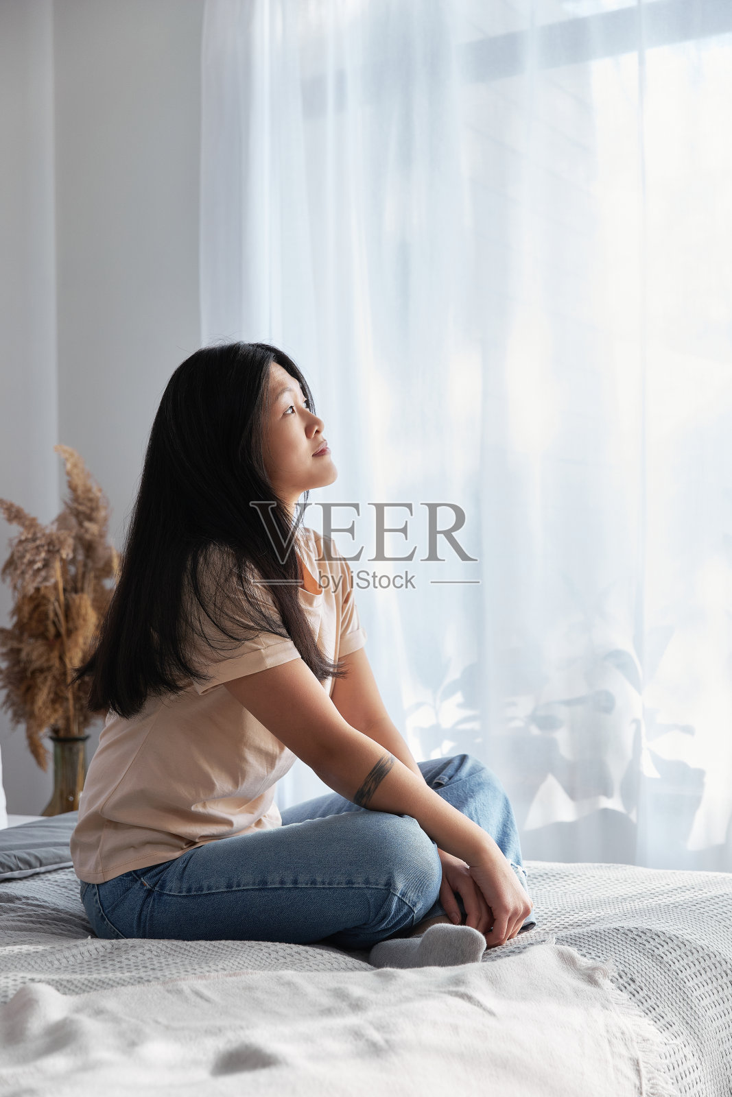 穿着休闲装的严肃亚洲女子坐在家里的床上，望着窗外照片摄影图片
