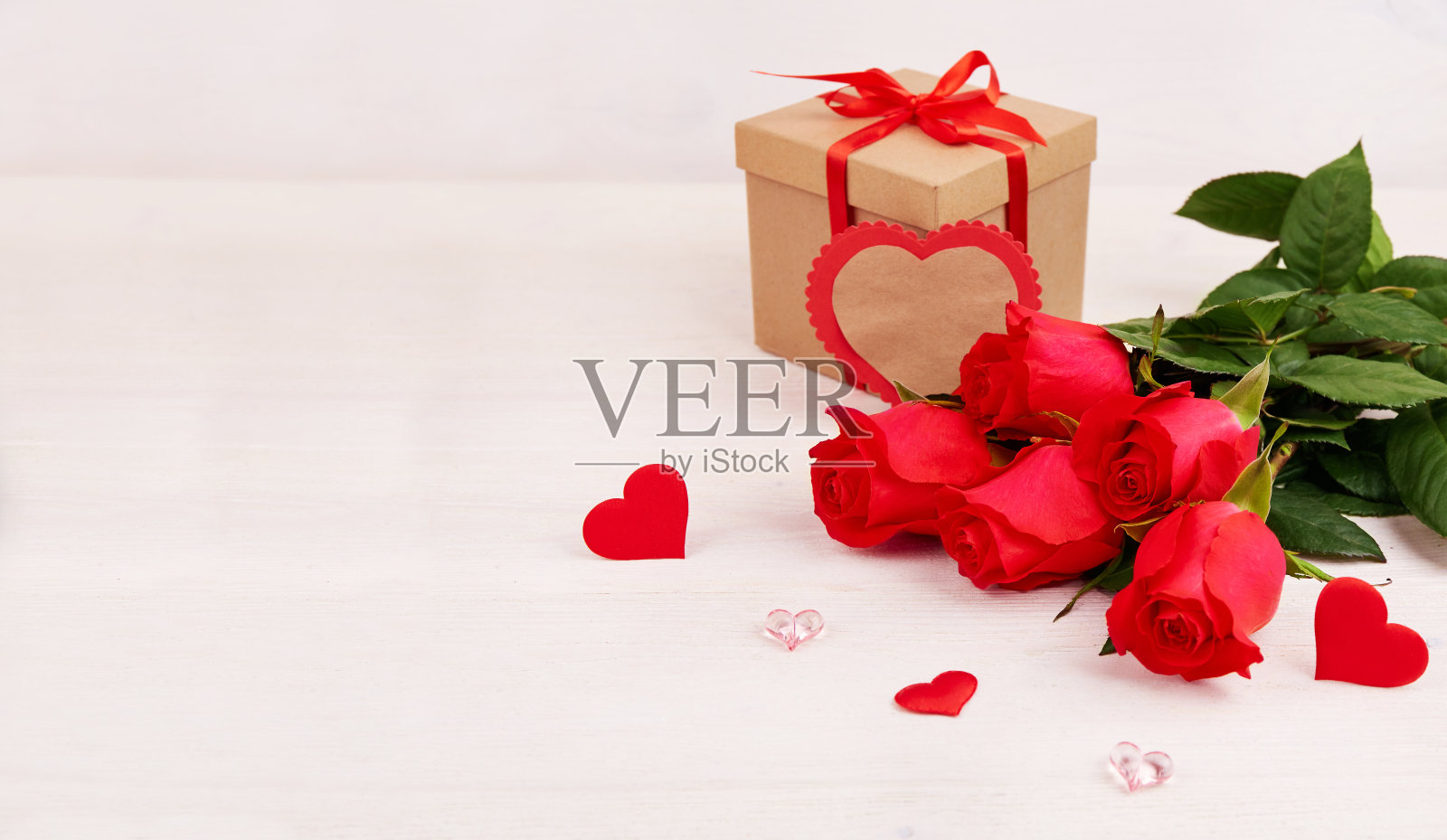红玫瑰花束，牛皮纸盒包装，搭配红丝带和心形装饰，置于白色木质背景上。情人节、母亲节、生日贺卡照片摄影图片