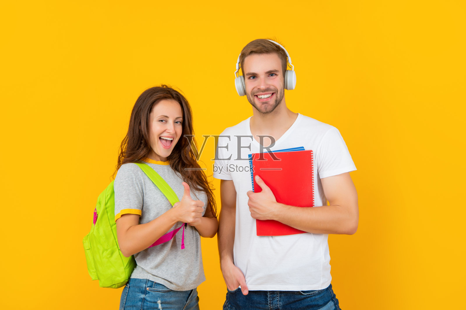 一对戴着耳机、背着练习册和背包的快乐学生们竖起大拇指表示:教育照片摄影图片