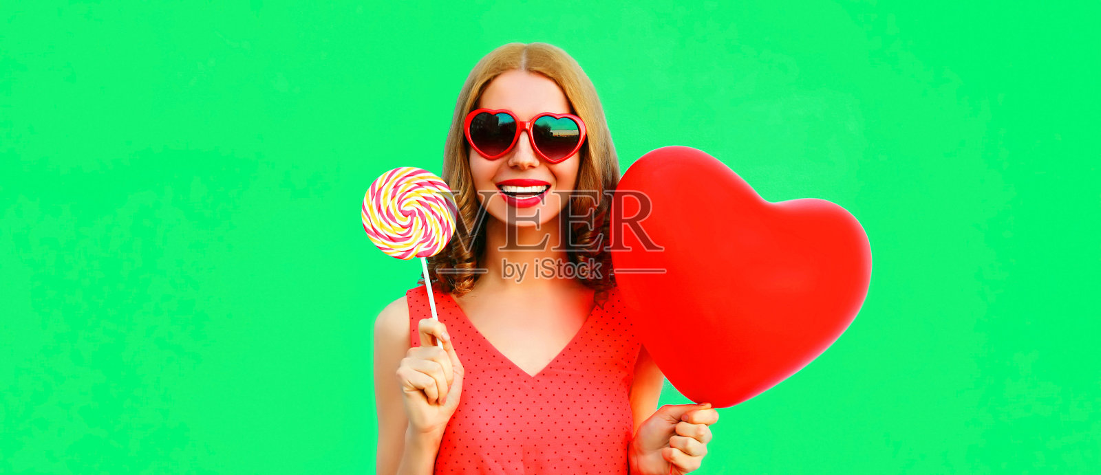 美丽快乐微笑的年轻女子，带着棒棒糖和一束红色的心形气球，戴着太阳镜在绿色的背景上照片摄影图片