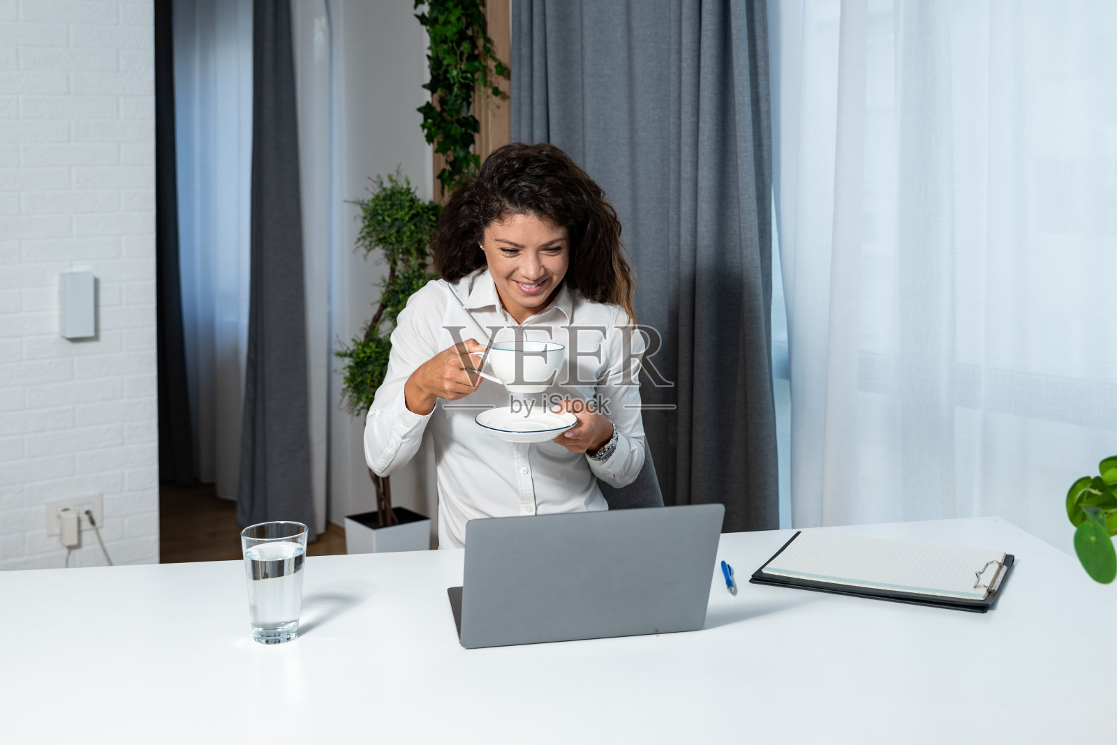 年轻快乐成功的女企业主在办公室用笔记本电脑工作时喝杯咖啡休息。女企业家，自信的女士，喝着热饮在电脑上阅读电子邮件照片摄影图片