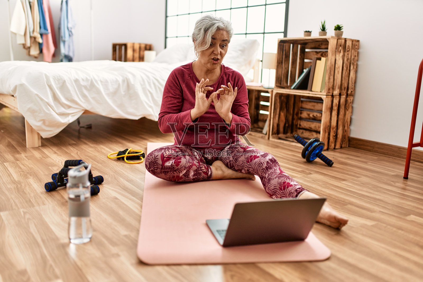 一个灰白头发的中年妇女坐在卧室的地板上进行视频通话训练。照片摄影图片