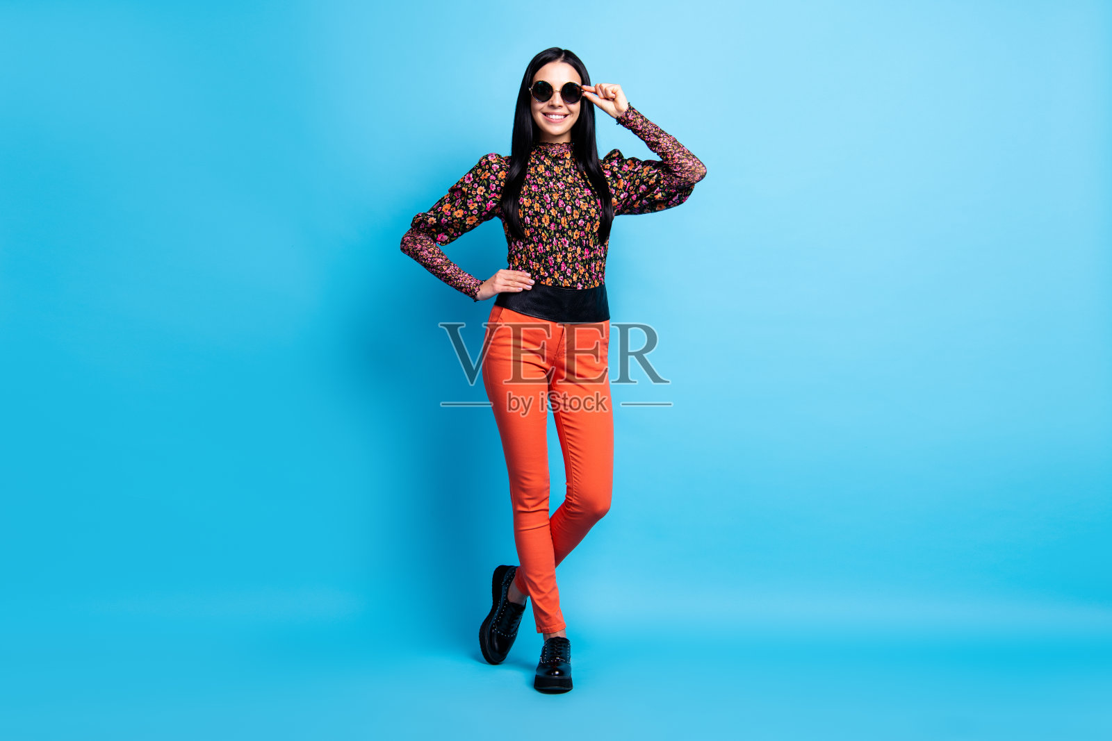 照片自信的女士摆姿势看相机穿复古眼镜花印花衬衫裤子靴子孤立的蓝色背景照片摄影图片