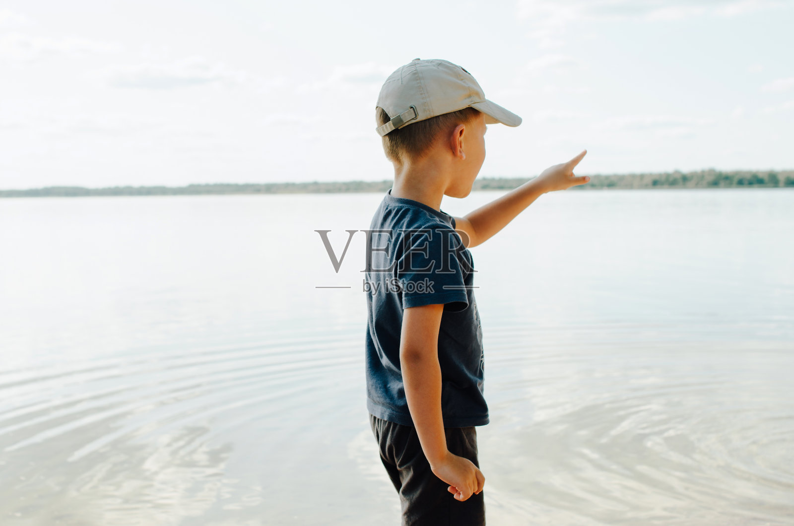 在阳光明媚的夏日户外，一个戴着帽子的白人男孩独自站在岸边的水边，在地平线上伸出手指。在大自然中，孩子看向别处。照片摄影图片