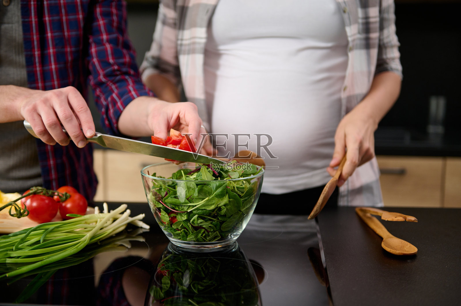 聚焦男性手持菜刀将番茄片放入碗中，背景是站在厨房岛台旁的怀孕妻子照片摄影图片