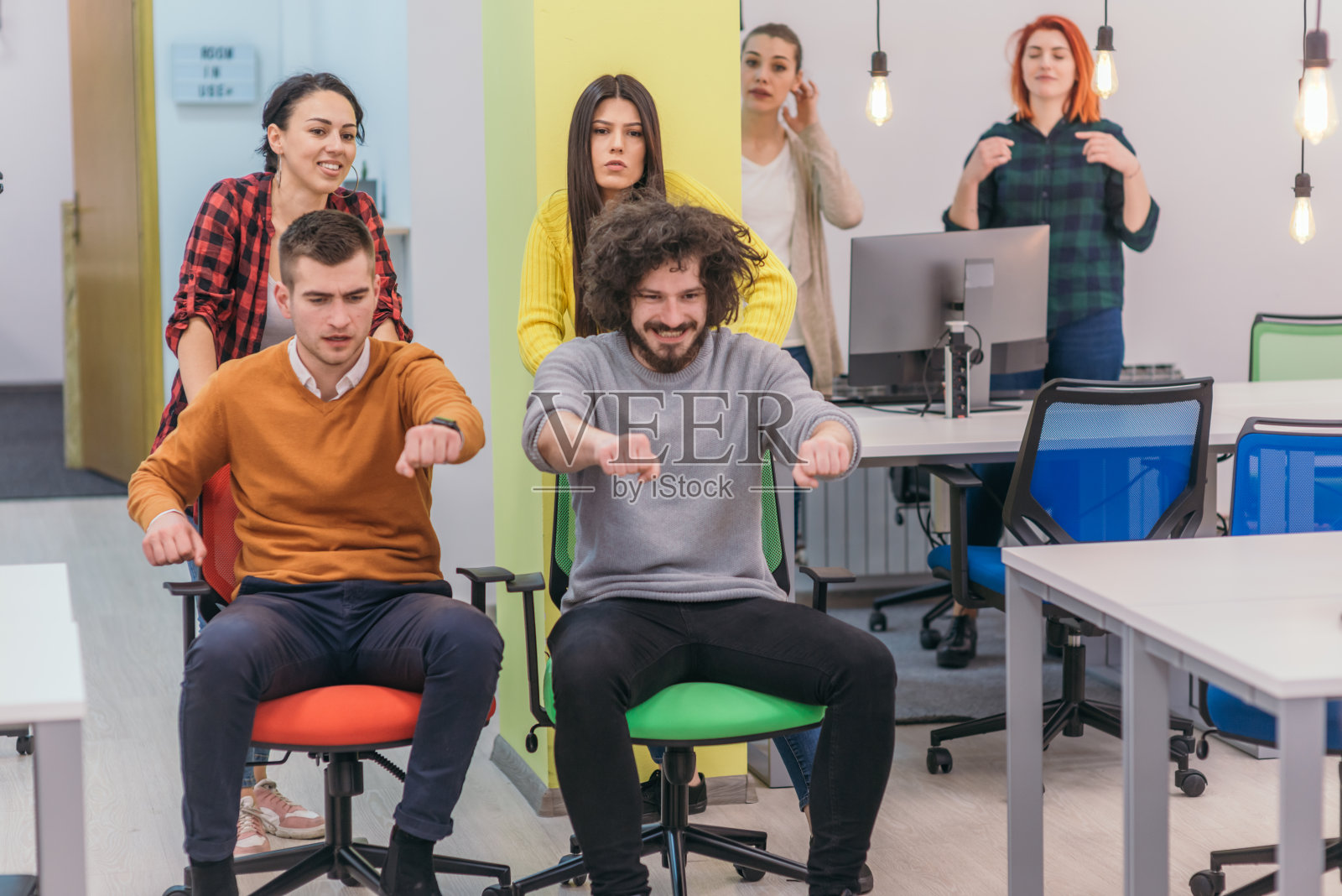 团队建设和办公室乐趣：四个年轻快乐的商务人士穿着休闲装，在办公椅上比赛并微笑照片摄影图片