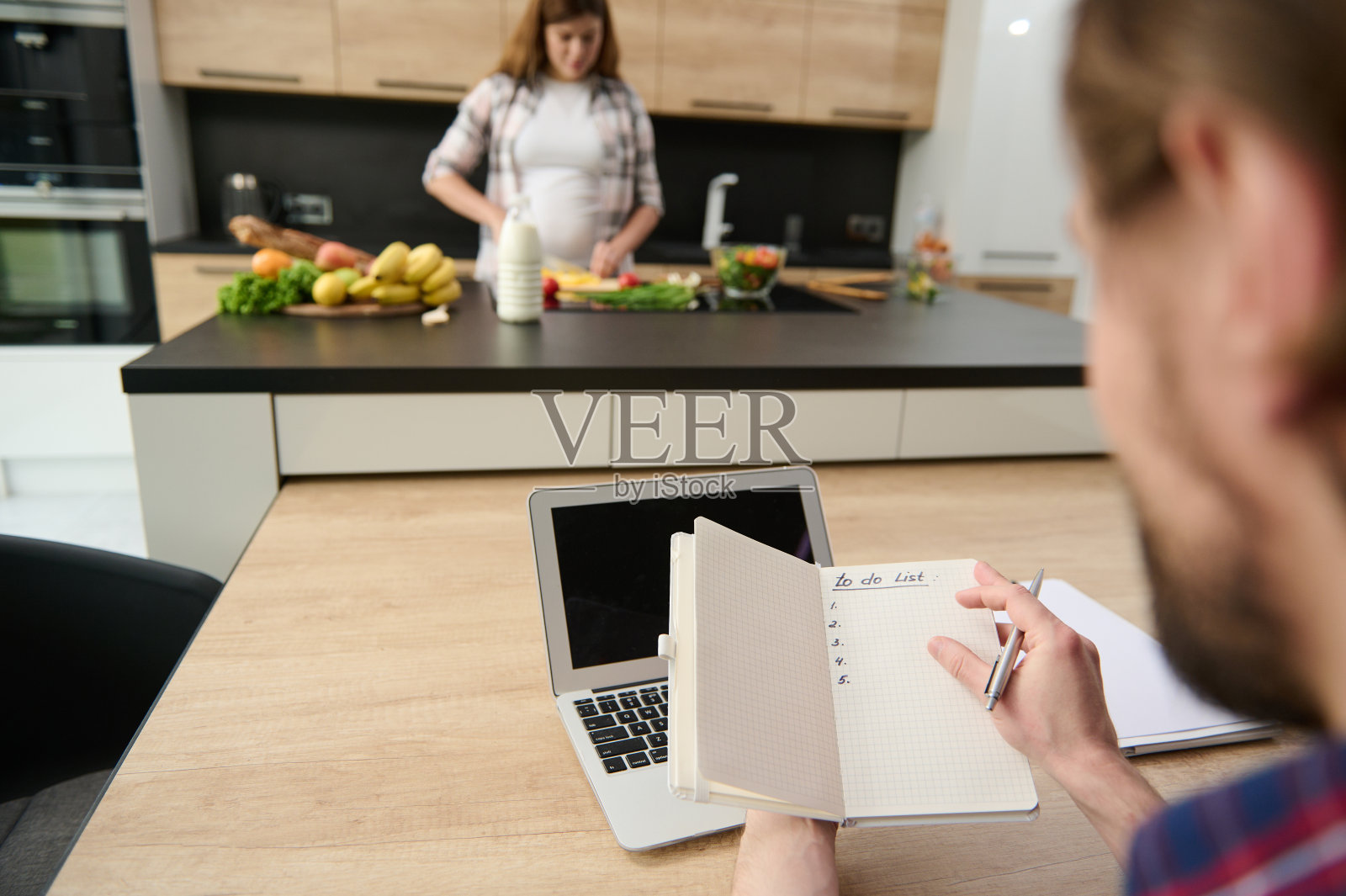 温柔的焦点在一个男人手里的笔记本电脑上工作，背景是他心不在焉的怀孕的妻子准备健康的食物在厨房岛。家庭生活的概念照片摄影图片