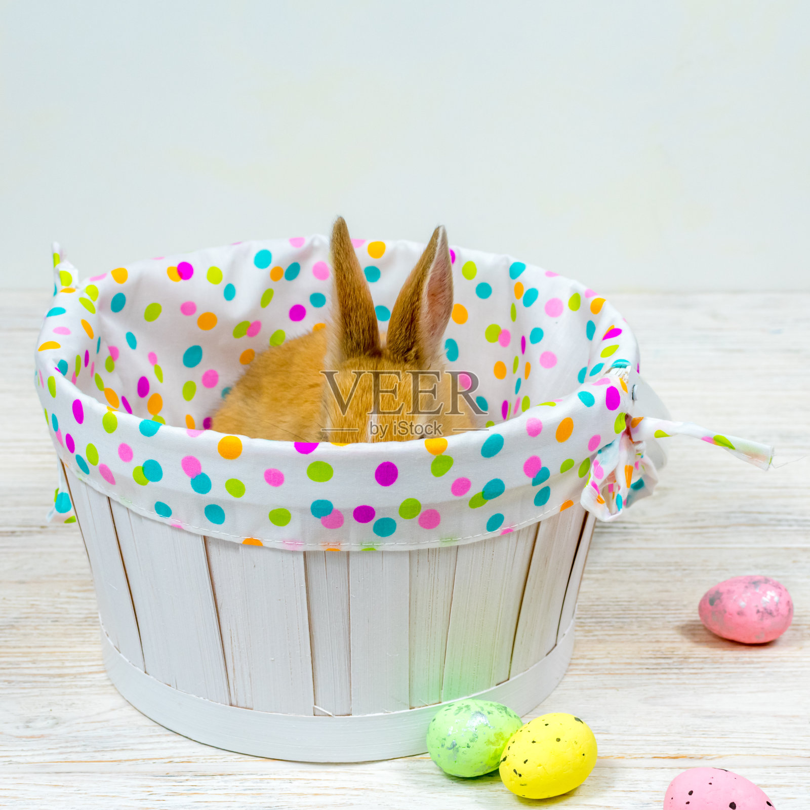 兔子耳朵从白色的篮子里伸出来，旁边是白色的桌子上的彩色复活节彩蛋。铭文的地方。复活节的概念照片摄影图片