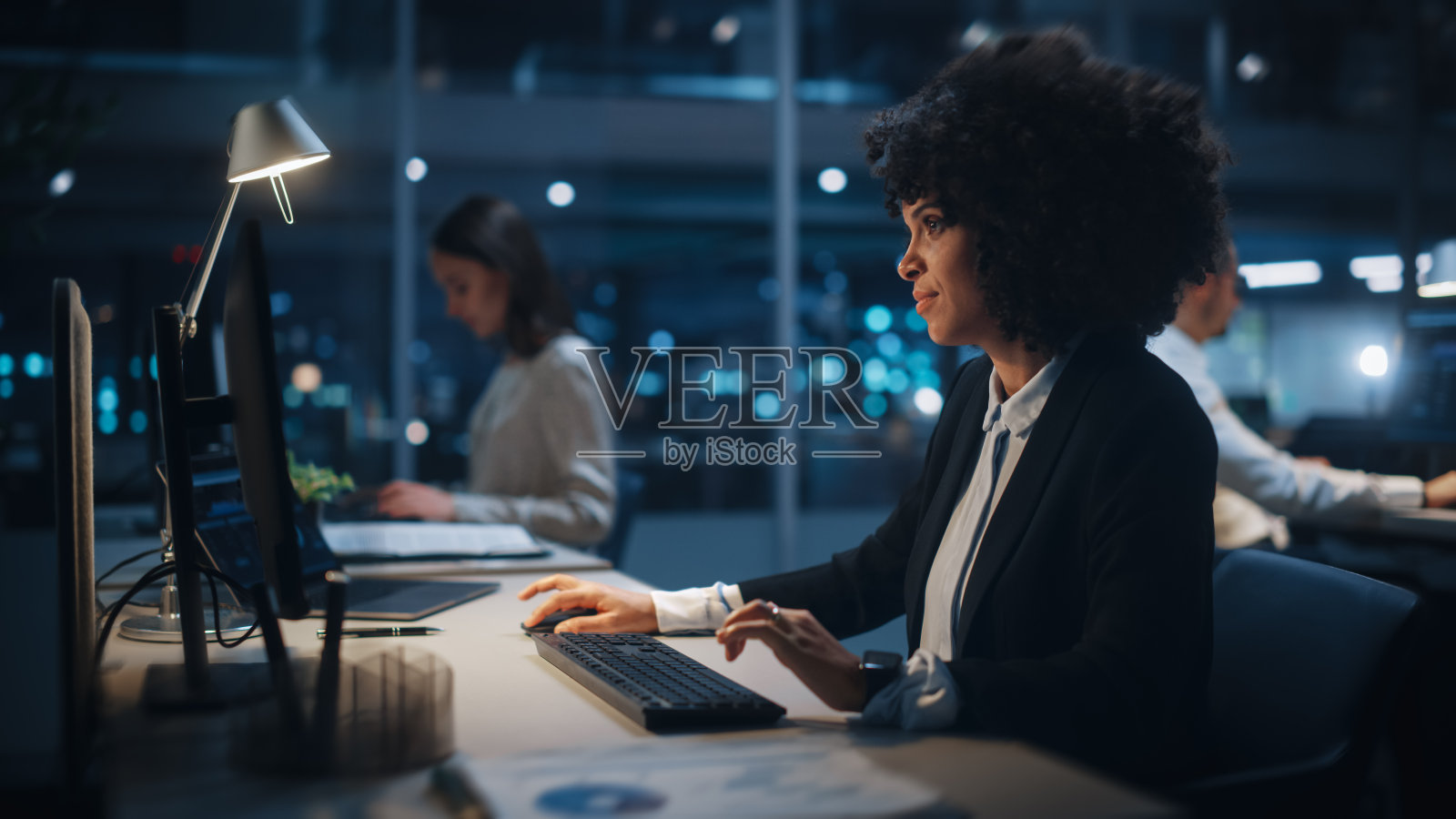 《晚上在大公司办公室:非裔美国女性经理使用电脑的肖像》，商业人士和专家在她身边工作，分析统计数据，商业数据，营销计划。照片摄影图片