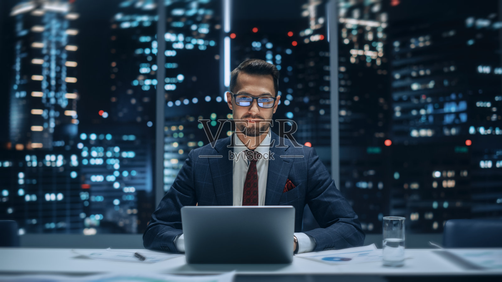 一个成功的英俊的商人的肖像，在大城市的办公室工作在笔记本电脑的深夜。快乐称职的经理在时尚的深蓝色西装微笑和相机摆姿势。照片摄影图片