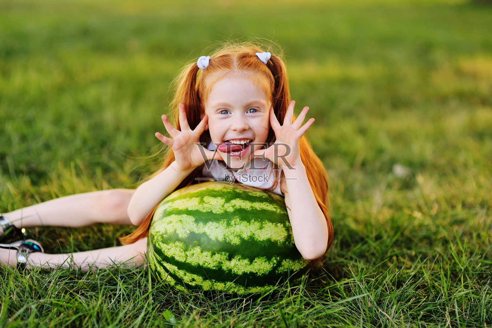 一个红发女孩，做着西瓜脸，微笑着坐在公园的草地上照片摄影图片