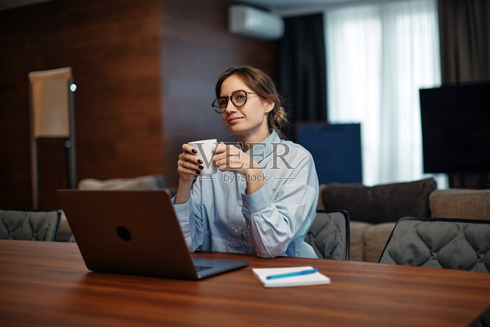 微笑的女员工坐在办公桌前，拿着笔记本电脑在家办公，拿着咖啡茶杯享受清晨，做梦。遥远的工作照片摄影图片