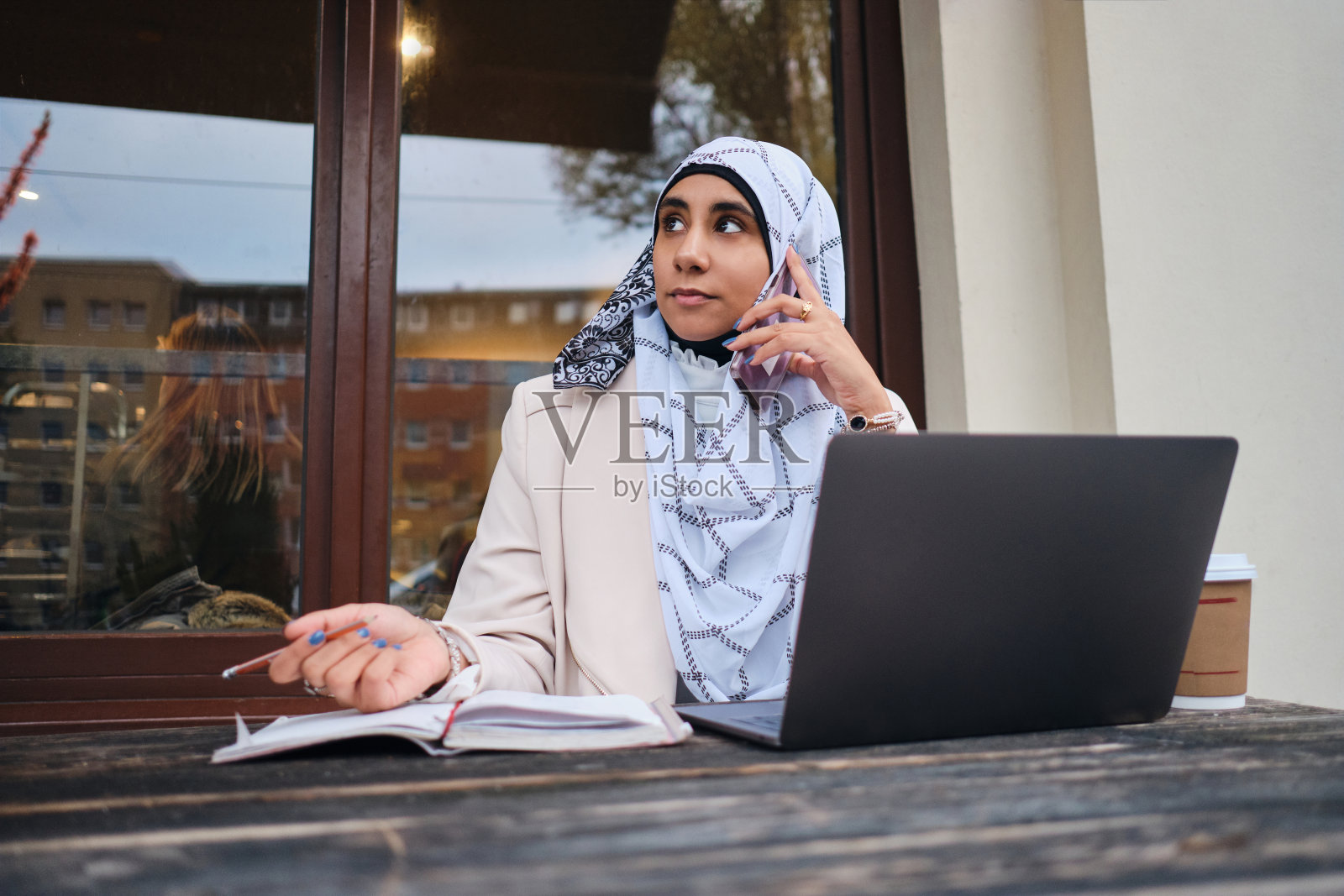 在城市的街道上，一位年轻的阿拉伯女商人戴着头巾，自信地用智能手机聊天，用笔记本电脑工作照片摄影图片