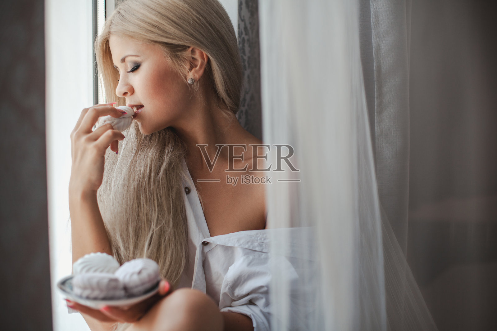 一个金发长发的美丽女孩独自站在窗前，吃着香甜的棉花糖照片摄影图片