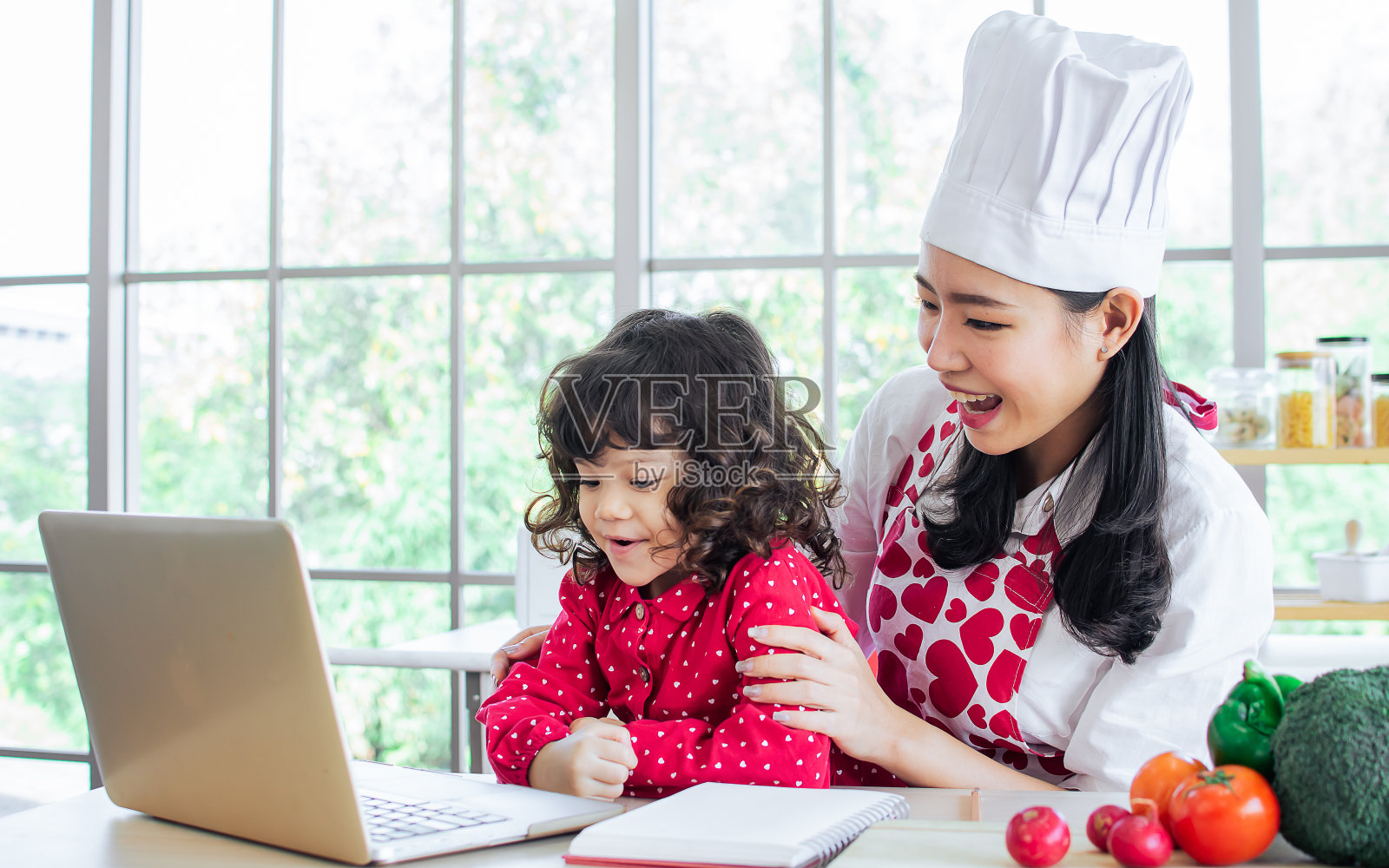 美丽的亚洲母亲煮早餐，同时照顾她的混血可爱的小女儿做作业，在线学习或学习，坐在厨房在家里，微笑着幸福。照片摄影图片