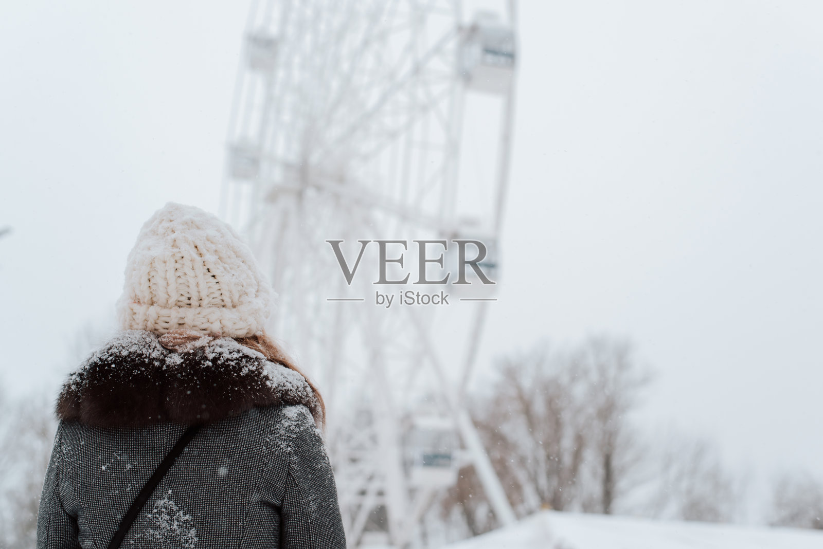 年轻女子的后视图，在冰雪覆盖的外套和针织白色帽子看摩天轮在冬季散步在公园，户外。本空间照片摄影图片