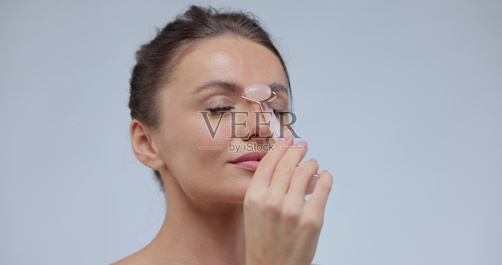 美丽的女人使用石头面部辊在灰色的背景。年轻的模特有着完美光滑的皮肤。照片摄影图片