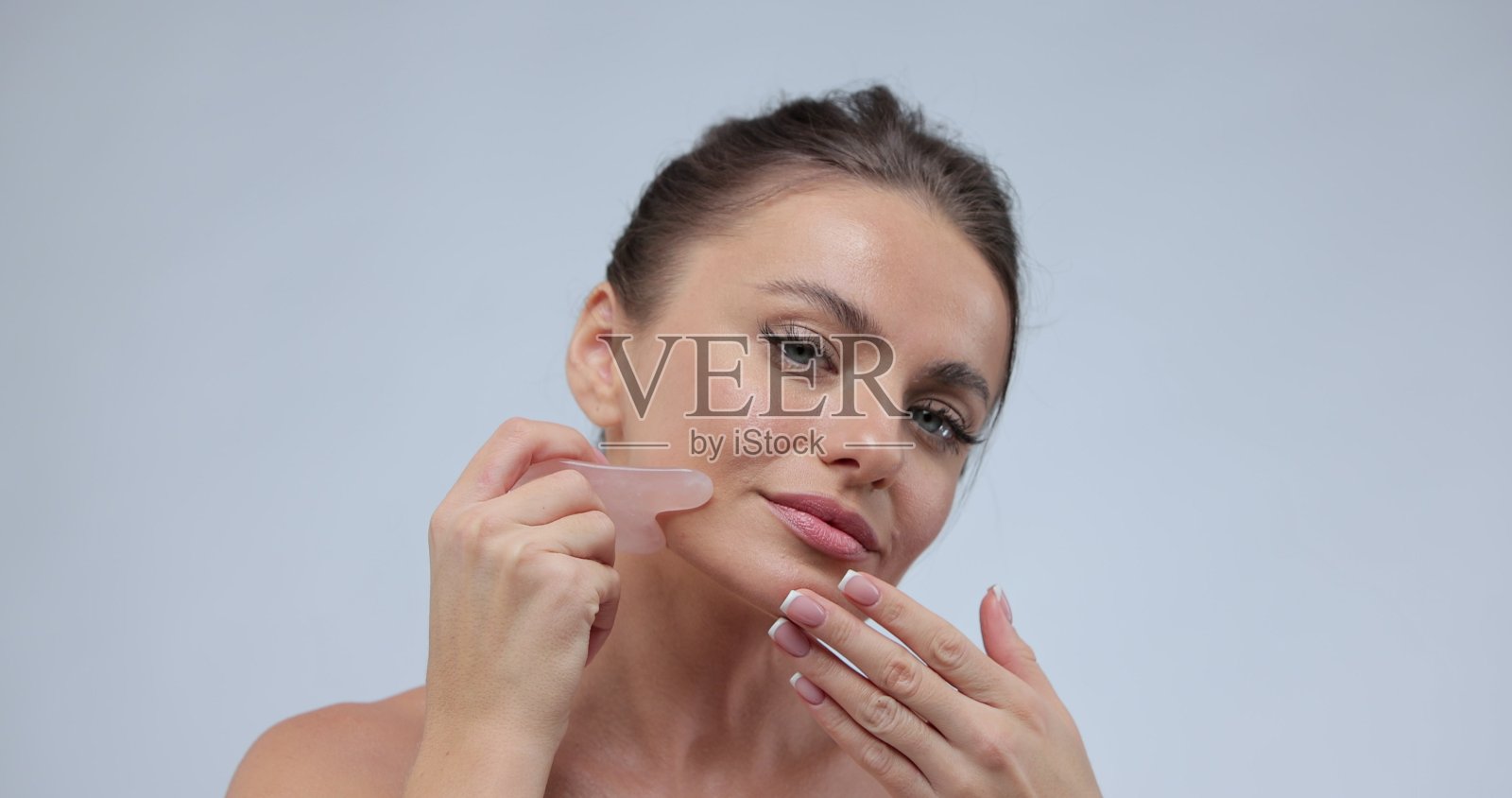 白种人年轻模特不化妆，一边看着相机一边用刮痧石做面部按摩。面部按摩。照片摄影图片