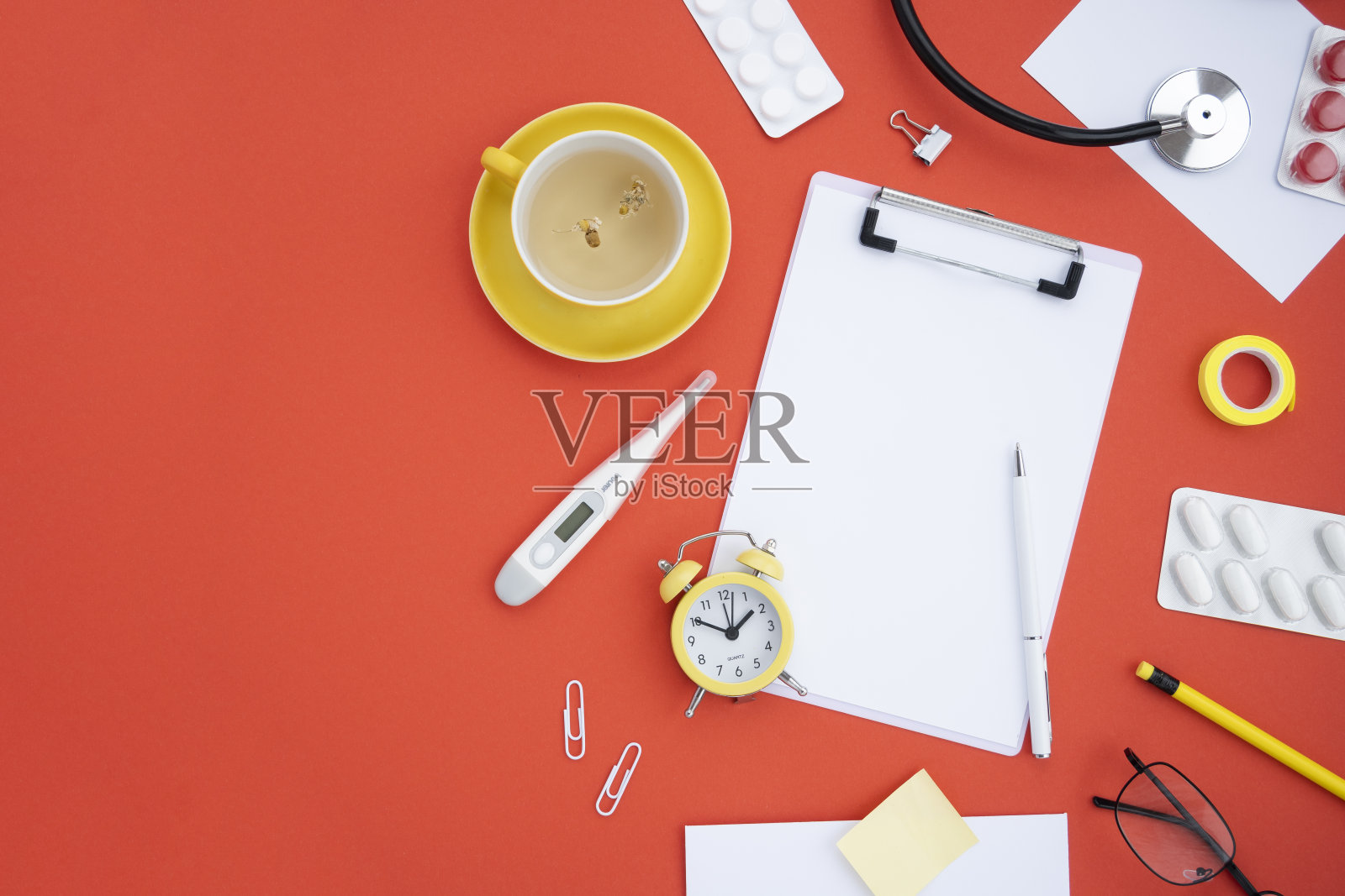 顶部视图的医生的桌面在红色背景与复制空间。用于笔记的药片，听诊器，眼镜，药丸，药物，一杯花草茶，手表。平坦的躺,模型照片摄影图片