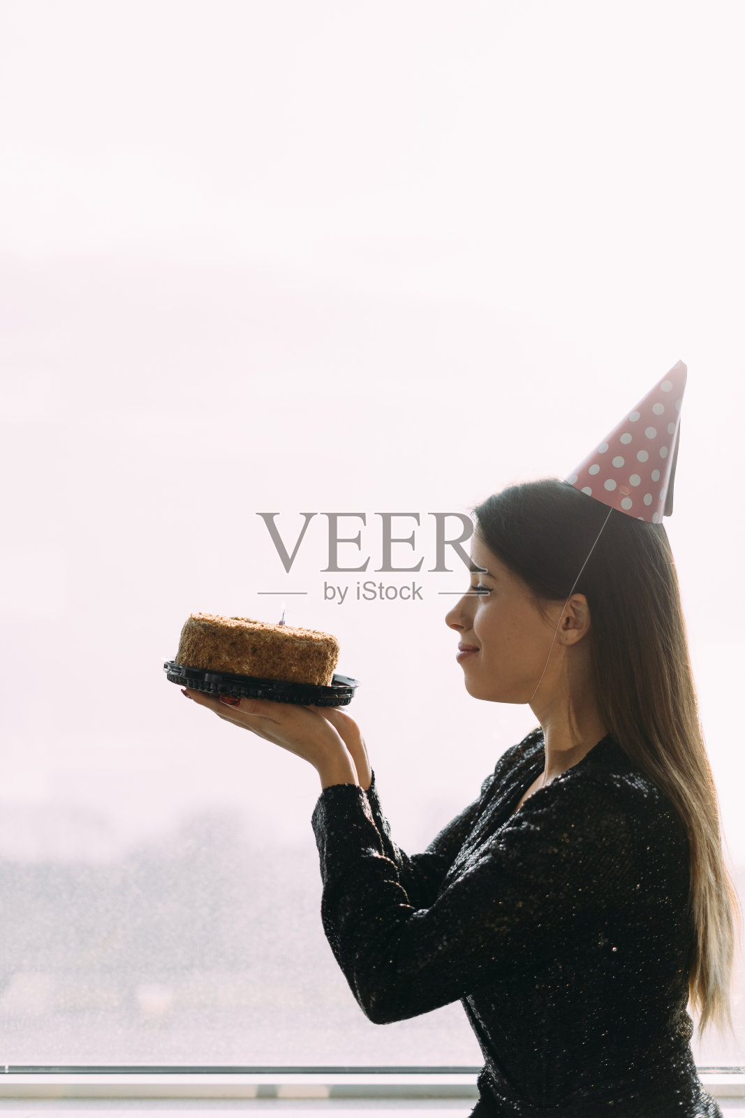 生日快乐，美丽的年轻女子拿着蛋糕看着蜡烛，站在办公室的窗户前，灯光背景，侧影观看照片摄影图片