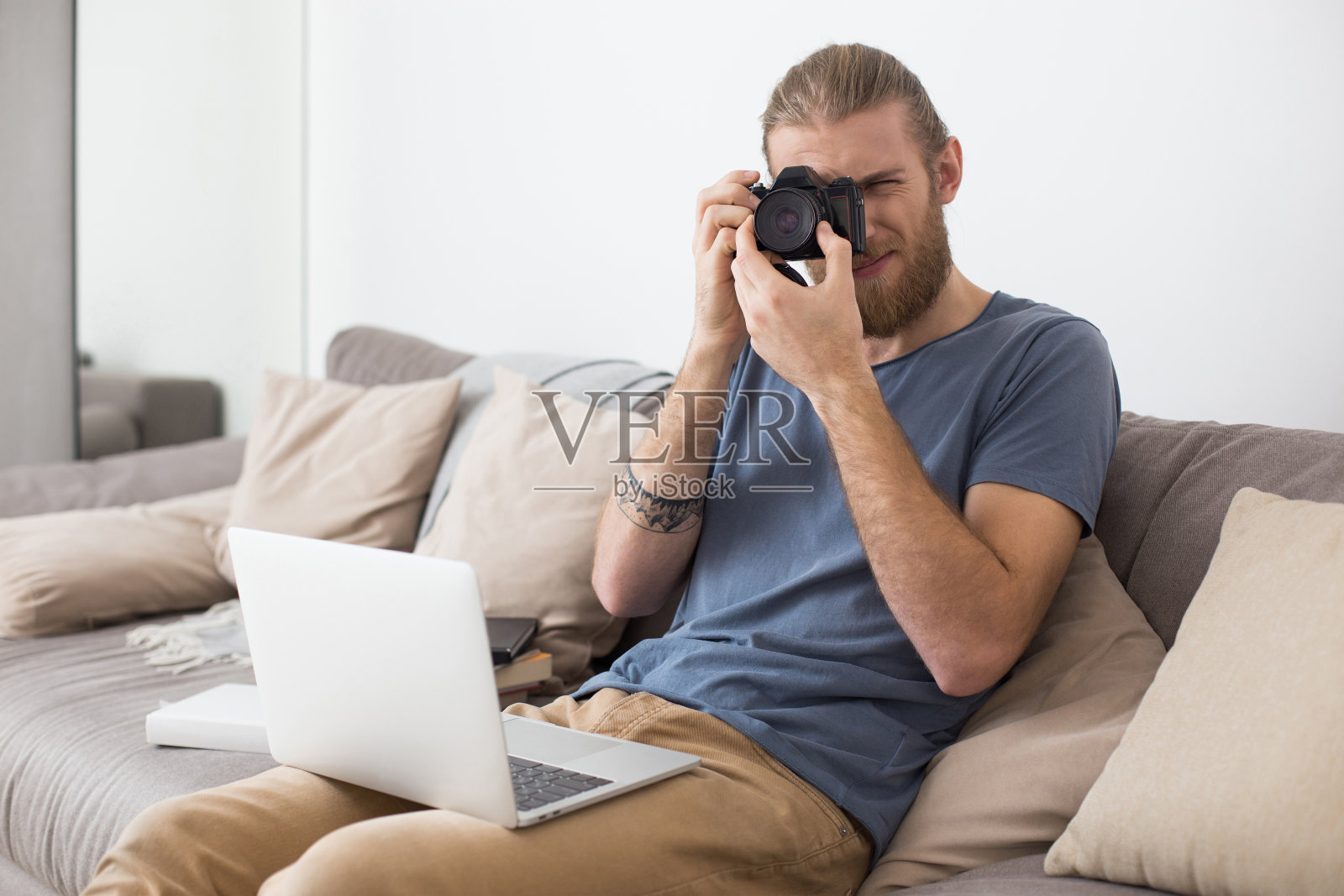 一个年轻人坐在灰色的沙发上，膝盖上放着笔记本电脑，手里拿着相机照片摄影图片