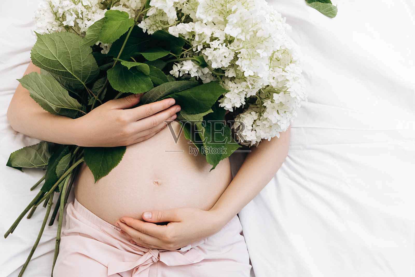 一个认不出来的孕妇的手轻轻抚摸着肚子躺在舒适的床上，捧着美丽新鲜的夏日鲜花。未来母亲的期望。副本的空间。母亲的概念照片摄影图片