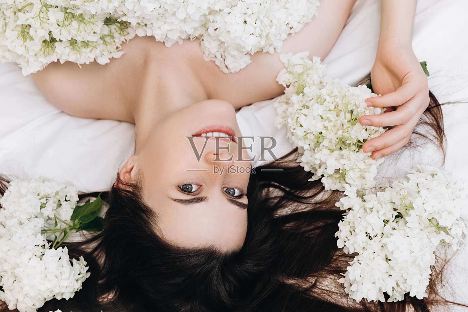 无忧无虑的早晨。近距离的肖像，欢快的年轻微笑的女人20岁放松在舒适的白色床在家里，积极地看着相机，享受新鲜的春天的花朵。人的情感概念照片摄影图片