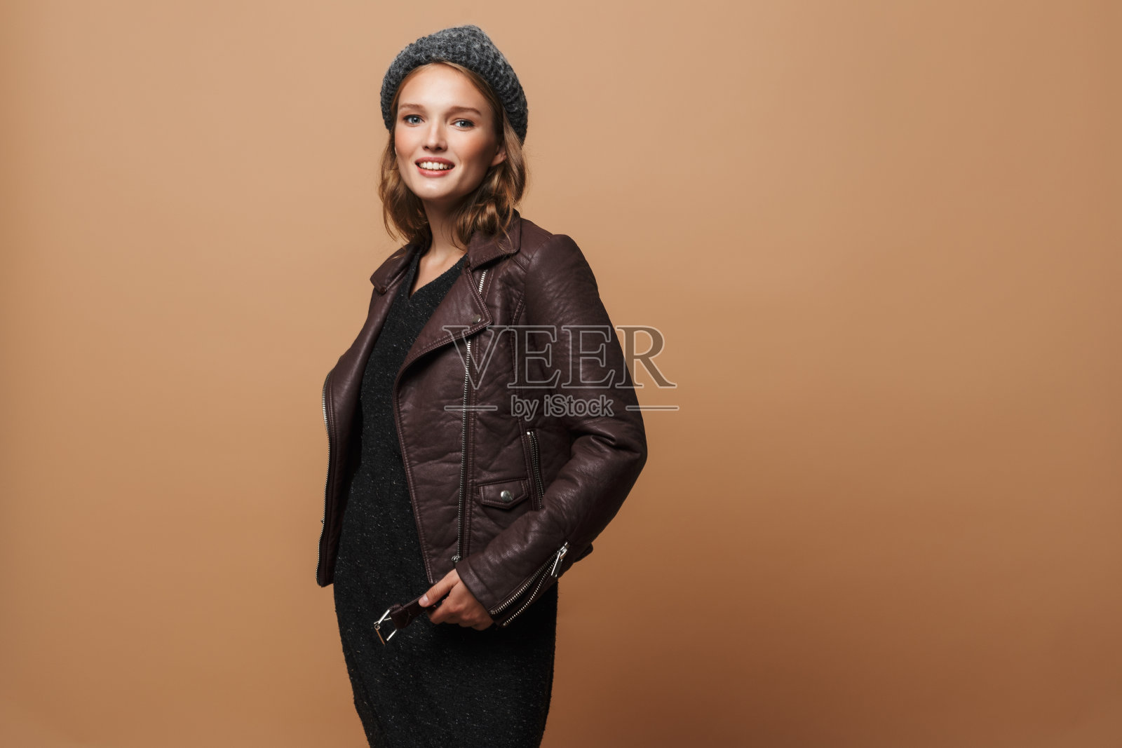 年轻漂亮的微笑的女人，在帽子，皮夹克和黑色连衣裙梦幻般的看着镜头在米色的背景照片摄影图片