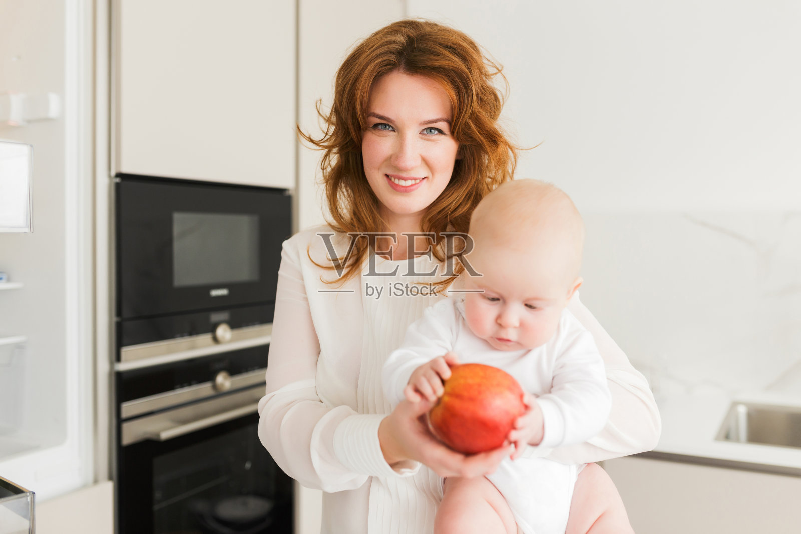 妈妈在厨房里面带微笑，抱着她可爱的小宝宝和大红苹果，开心地看着镜头照片摄影图片