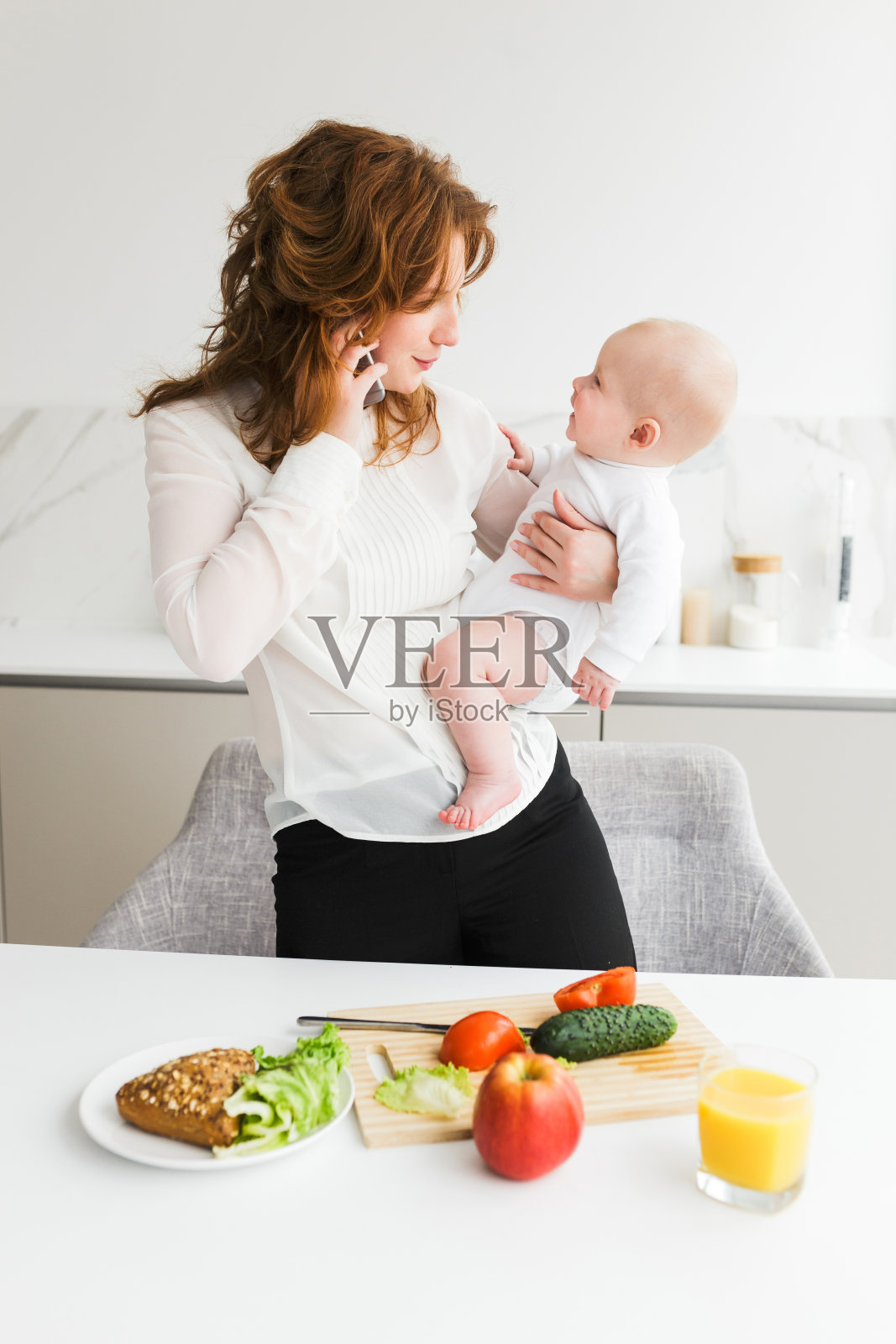美丽的母亲抱着她快乐的小婴儿，一边打电话，一边在厨房做饭照片摄影图片
